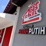 Public cooperative building with bold red-and-white Merah Putih sign mounted on gray wall; red doors along a white walkway. A person in a hijab approaches one door.