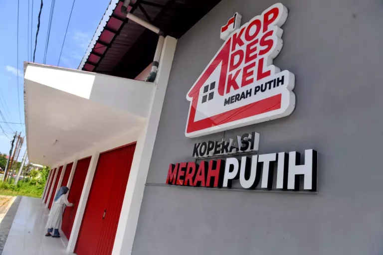 Public cooperative building with bold red-and-white Merah Putih sign mounted on gray wall; red doors along a white walkway. A person in a hijab approaches one door.