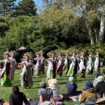Dancers in white tops and colorful skirts perform a line dance on a grassy stage while a seated audience watches outdoors under bright sun.