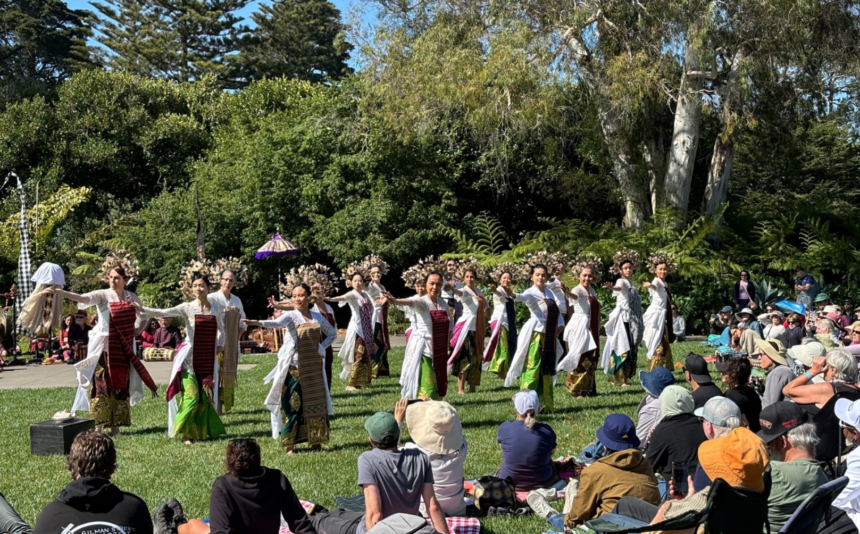 Dancers in white tops and colorful skirts perform a line dance on a grassy stage while a seated audience watches outdoors under bright sun.