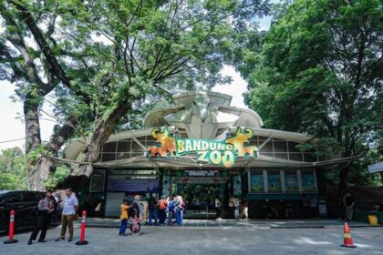 Bandung Zoo entrance with green sign and two orange animal figures, visitors entering on a sunny day under leafy trees