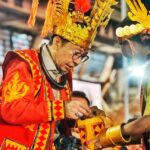 Two men in bright ceremonial costumes and tall gold headdresses exchange a gift at a festive event.