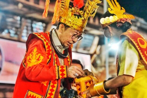 Two men in bright ceremonial costumes and tall gold headdresses exchange a gift at a festive event.