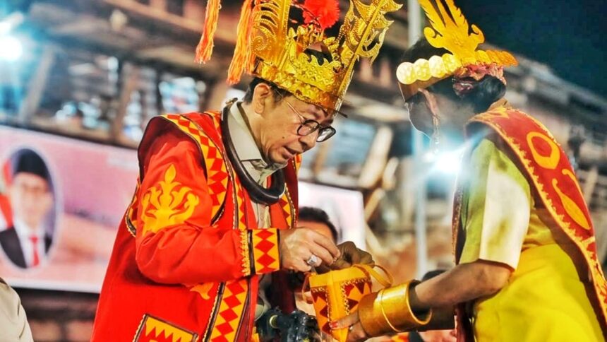 Two men in bright ceremonial costumes and tall gold headdresses exchange a gift at a festive event.