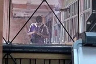 Two people standing close together inside a glass-walled hallway, one wearing a purple shirt, looking down at a phone.