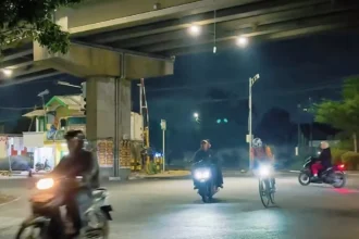 Night street scene under an overpass with several motorcyclists and cyclists riding toward the camera; headlights illuminate the road at dusk.