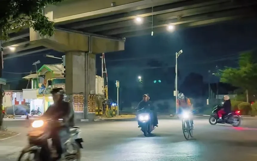 Night street scene under an overpass with several motorcyclists and cyclists riding toward the camera; headlights illuminate the road at dusk.