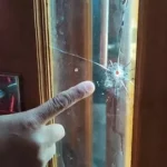 A hand points at a spider crack in the glass panel of a wooden door, highlighting the damage.