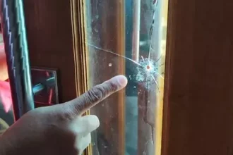 A hand points at a spider crack in the glass panel of a wooden door, highlighting the damage.