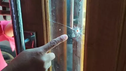 A hand points at a spider crack in the glass panel of a wooden door, highlighting the damage.