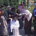 Group of women in batik and hijabs gather around a seated girl as they adjust her white outfit at an outdoor ceremony of cultural significance.