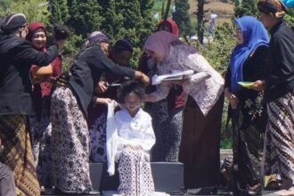 Group of women in batik and hijabs gather around a seated girl as they adjust her white outfit at an outdoor ceremony of cultural significance.