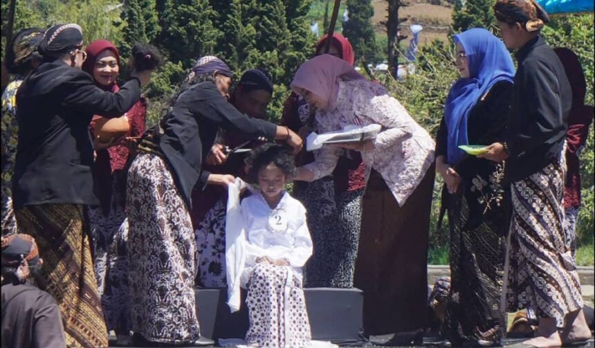 Group of women in batik and hijabs gather around a seated girl as they adjust her white outfit at an outdoor ceremony of cultural significance.