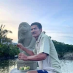 Man sitting beside a stone monkey statue by a waterway at sunset, holding a drink with a bowl of fruit nearby.