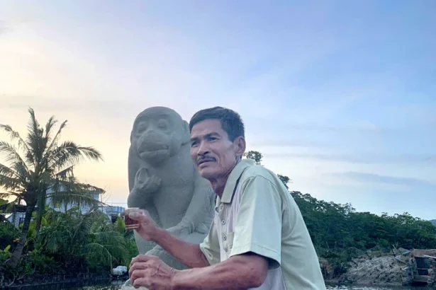 Man sitting beside a stone monkey statue by a waterway at sunset, holding a drink with a bowl of fruit nearby.