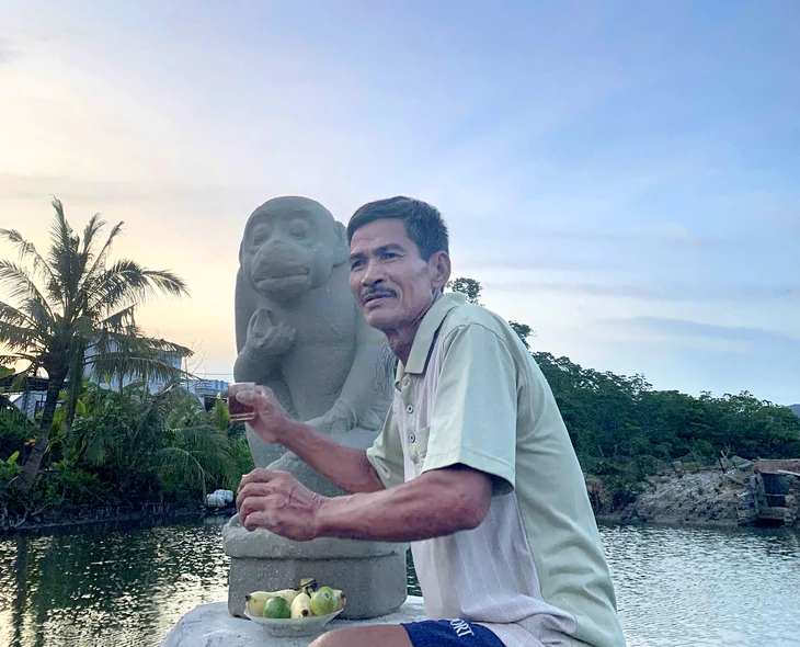 Man sitting beside a stone monkey statue by a waterway at sunset, holding a drink with a bowl of fruit nearby.