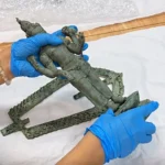 Gloved hands join pieces of a green bronze statue on a white work surface during restoration.
