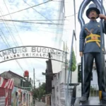 Archway entrance sign reading 'KAMPUNG BUCIS' with 'JALAN SRI KALOM' visible; a tall guard statue in uniform stands beside it holding a spear.
