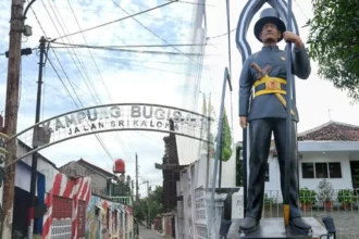 Archway entrance sign reading 'KAMPUNG BUCIS' with 'JALAN SRI KALOM' visible; a tall guard statue in uniform stands beside it holding a spear.