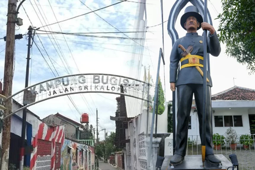 Archway entrance sign reading 'KAMPUNG BUCIS' with 'JALAN SRI KALOM' visible; a tall guard statue in uniform stands beside it holding a spear.