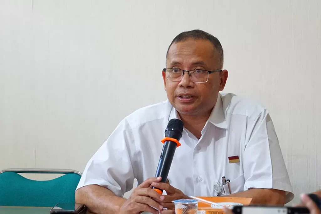 Man in a white shirt and glasses speaks into a microphone at a conference table.