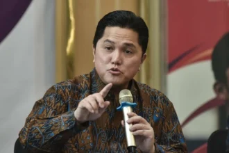 Man in a brown batik shirt speaks into a gold handheld microphone, gesturing with his index finger as he makes a point during a discussion.
