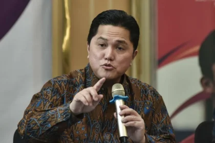 Man in a brown batik shirt speaks into a gold handheld microphone, gesturing with his index finger as he makes a point during a discussion.