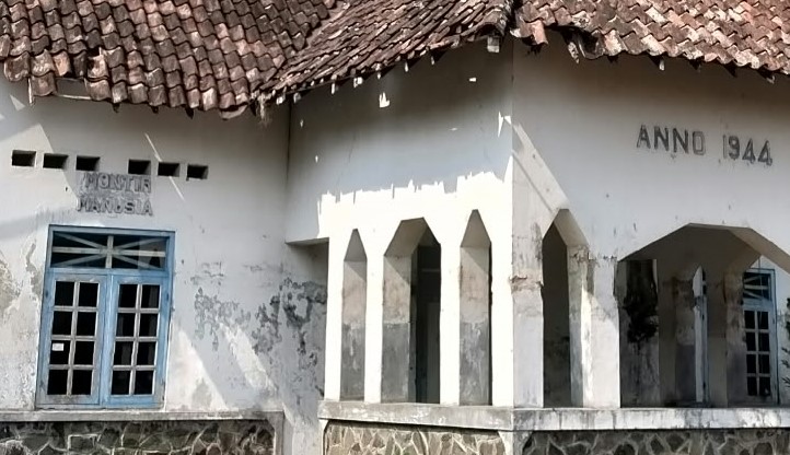 White weathered building with a blue-framed window, arched colonnade, and 'Anno 1944' carved on the wall, tiled roof in the background.