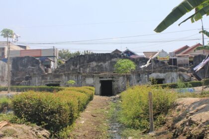 Old ruined concrete wall with overgrown plants and a narrow canal in front, in a tropical village with houses and power lines in the background.