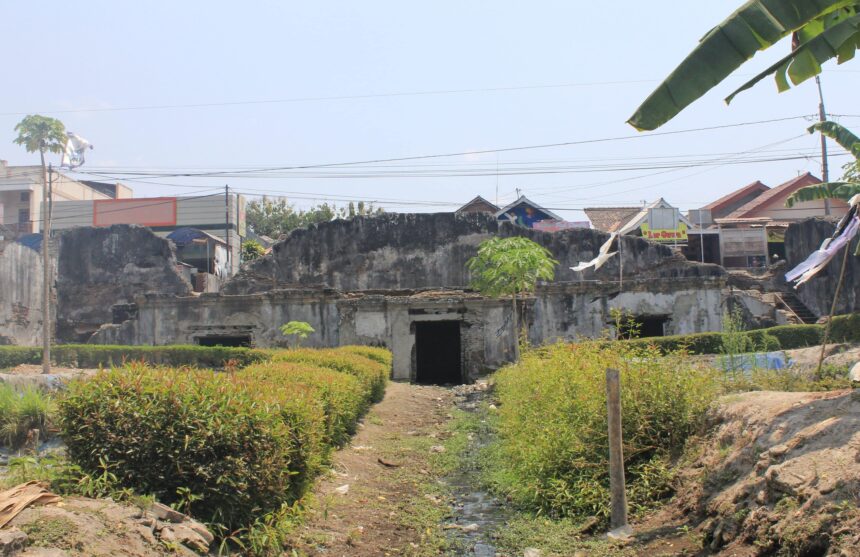 1bb00656208bb3329b58f8a012d07ffa - Mabur.co Old ruined concrete wall with overgrown plants and a narrow canal in front, in a tropical village with houses and power lines in the background.