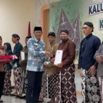 Group of people in traditional clothing on a stage, a man hands a certificate to another while others hold gifts, with a banner reading Kalurahan in the background.