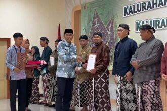 Group of people in traditional clothing on a stage, a man hands a certificate to another while others hold gifts, with a banner reading Kalurahan in the background.