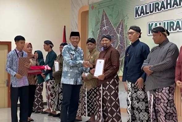 Group of people in traditional clothing on a stage, a man hands a certificate to another while others hold gifts, with a banner reading Kalurahan in the background.