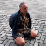 Man kneeling on a gray cobblestone surface, wearing a navy jacket and a brown batik shirt, looking to the right.