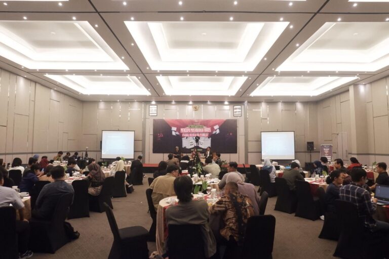 Stage with panel of speakers and a maroon banner at the front; attendees seated at round banquet tables in a well-lit conference hall.