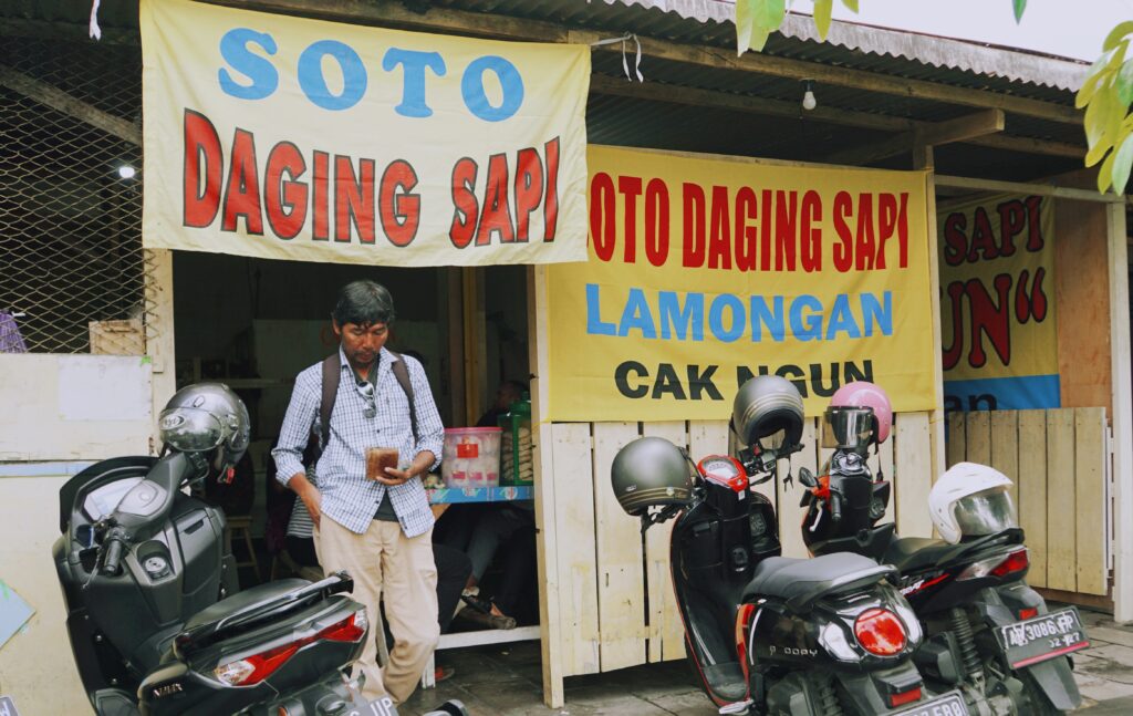 Storefront with large Indonesian banners advertising beef (daging sapi) and the town Lamongan; man stands at entrance amid parked motorbikes.