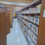 Library aisle in a library with tall wooden shelves filled with colorful books, white tiled floor, and overhead fluorescent lights; signs indicate general reading section.