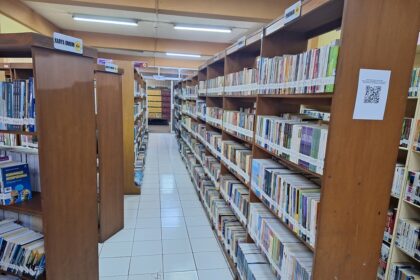 Library aisle in a library with tall wooden shelves filled with colorful books, white tiled floor, and overhead fluorescent lights; signs indicate general reading section.
