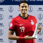 Soccer player in a red jersey with number 19, smiling, holding a silver trophy on a backdrop covered with Hyundai Cup logos.