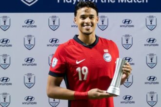 Soccer player in a red jersey with number 19, smiling, holding a silver trophy on a backdrop covered with Hyundai Cup logos.