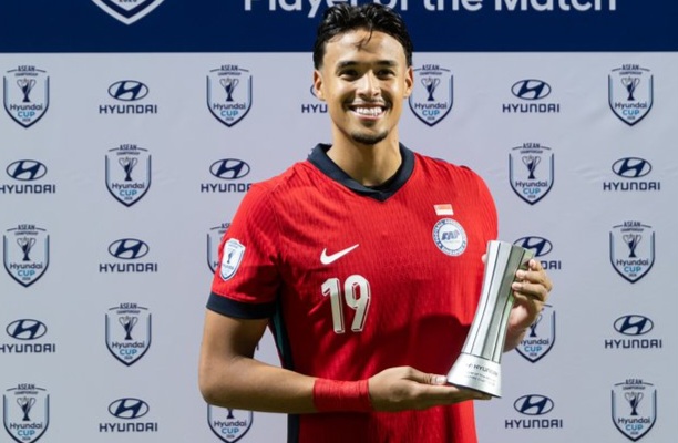 Soccer player in a red jersey with number 19, smiling, holding a silver trophy on a backdrop covered with Hyundai Cup logos.