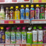 Row of colorful bottled drinks on supermarket shelf, with yellow, blue, and green caps and various fruit-tea and juice labels.