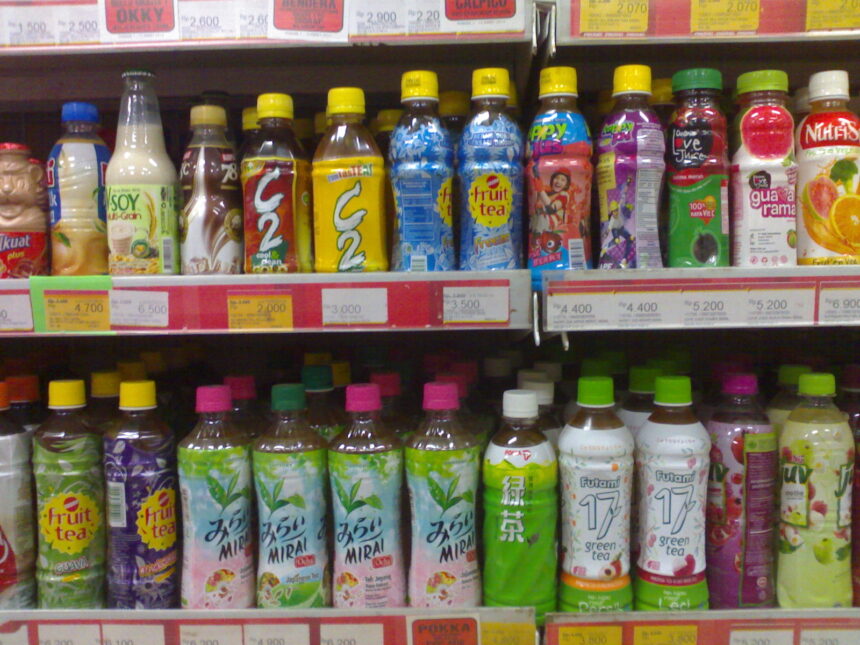 Row of colorful bottled drinks on supermarket shelf, with yellow, blue, and green caps and various fruit-tea and juice labels.