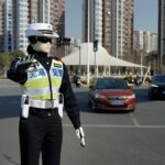 Traffic police officer in a reflective vest directing traffic at a busy urban intersection with tall buildings in the background.