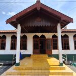 Front view of a white building with green base, yellow steps, and a large wooden gabled roof supported by white columns; arched windows line the facade under the roof.