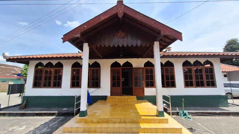Front view of a white building with green base, yellow steps, and a large wooden gabled roof supported by white columns; arched windows line the facade under the roof.