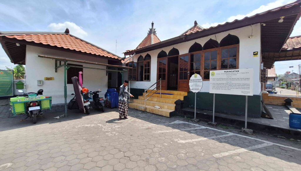 20260508_112527 - Mabur.co A small white building with arched dark windows and a red tile roof; a man in traditional clothing walks on a stone-paved courtyard beside parked motorcycles and a green cart right of center.