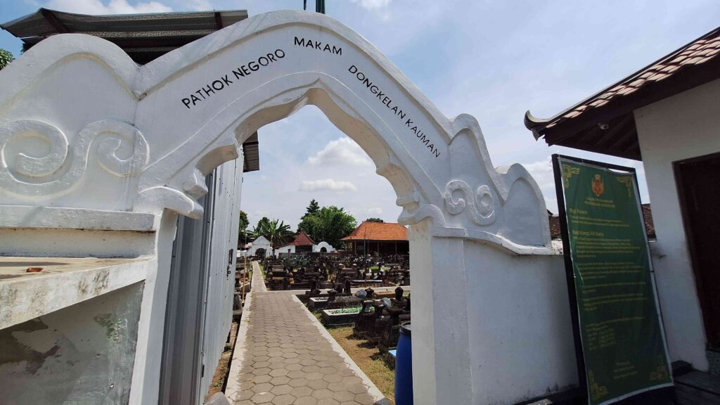White decorative arch marks the entrance to a cemetery, with a stone-paved path and rows of grave markers beyond it.