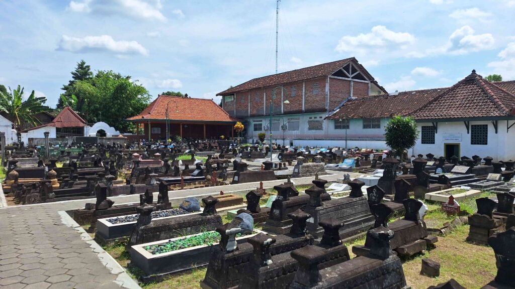 Wide view of a cemetery with many tombstones arranged in rows, a pavilion with a red-tiled roof, and brick buildings in the background under a blue sky.