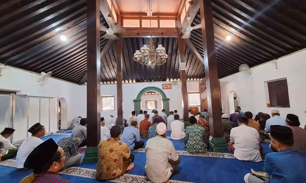 20260508_114805 - Mabur.co Muslim men praying inside a wooden-ceiling mosque, sitting on blue prayer carpets under a chandelier